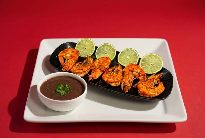Grilled shrimp with lime slices on a black plate, next to a bowl of dark sauce, on a white platter.