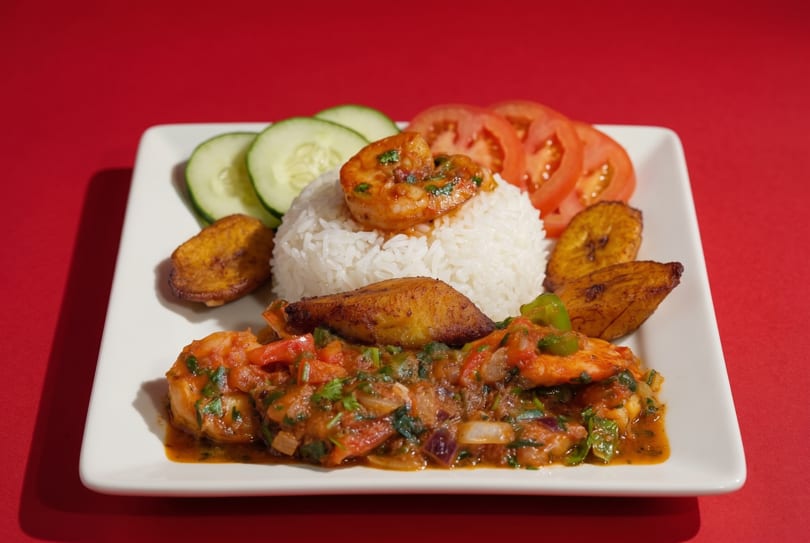 Shrimp creole with white rice, fried plantains, sliced cucumbers, and tomatoes on a white plate.