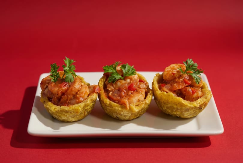 Three shrimp tostones with red sauce and parsley on a white rectangular plate against a red background.