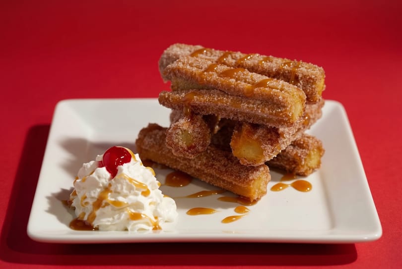Churros with caramel drizzle and whipped cream topped with a cherry on a white plate.